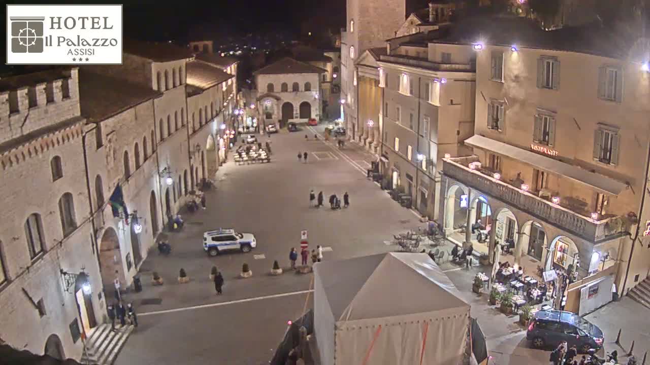 A nighttime, high-angle view of a town square in a European-style town, showing several buildings lit up, a few people walking around, and some cars parked.