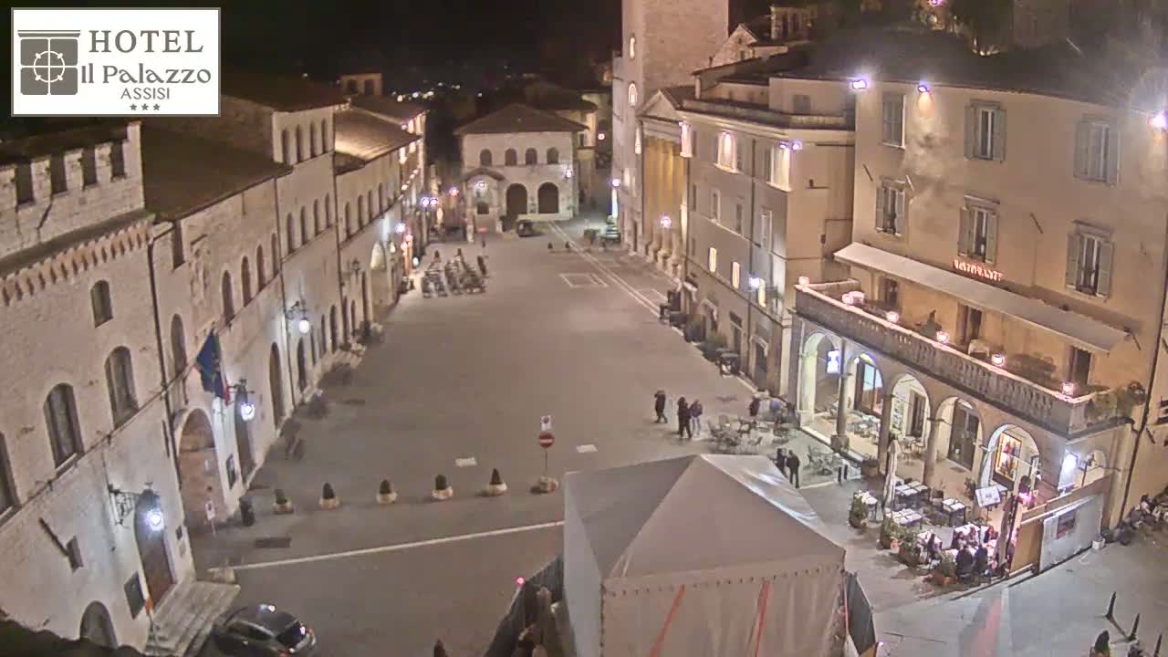 A nighttime view of a town square in Italy, with several buildings lit up and a few people walking around.