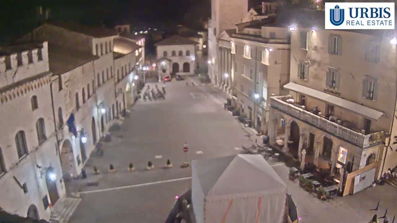 An empty town square at night, surrounded by old stone buildings, is illuminated by streetlights.