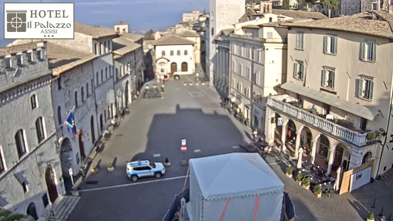 A mostly empty town square in a European town, under a sunny sky, is surrounded by several multi-story buildings, with a police car and a large white tent visible in the square.
