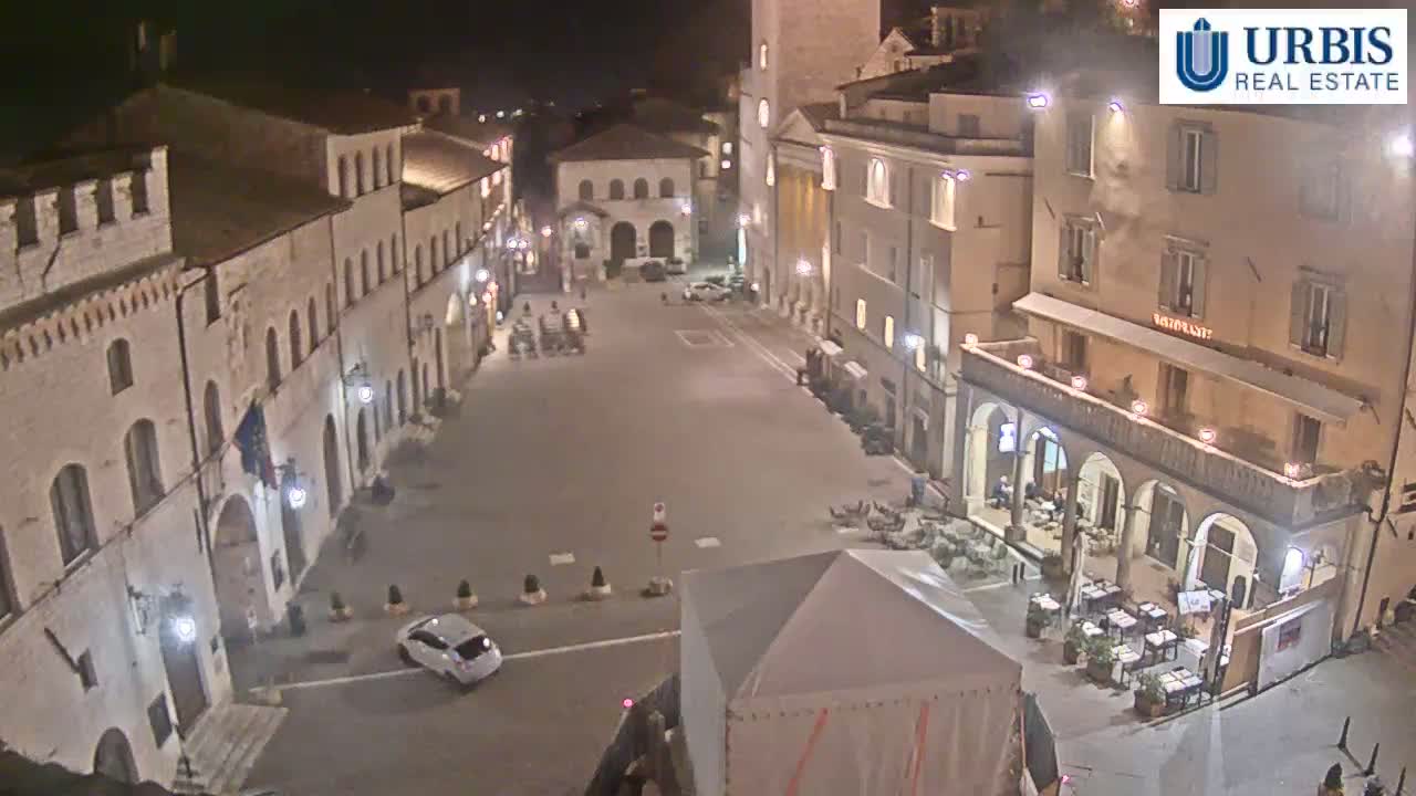A nighttime view of a town square with several buildings illuminated, a car parked in the square, and outdoor seating at a restaurant.