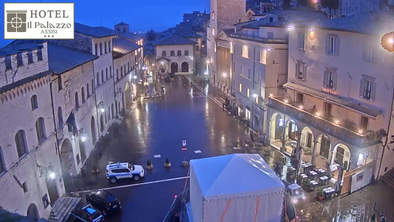 A rain-slicked town square at night, surrounded by illuminated stone buildings, with a few cars and a temporary tent visible.