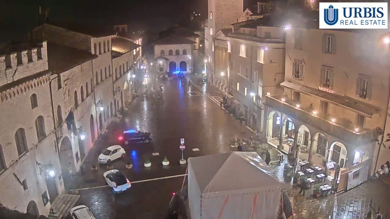 A rain-slicked town square at night, surrounded by illuminated buildings, with several cars parked and a temporary structure in the foreground.
