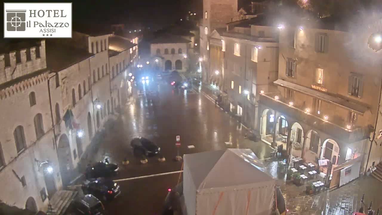 A rain-slicked town square at night, surrounded by illuminated buildings, with a few cars parked and some outdoor seating visible.