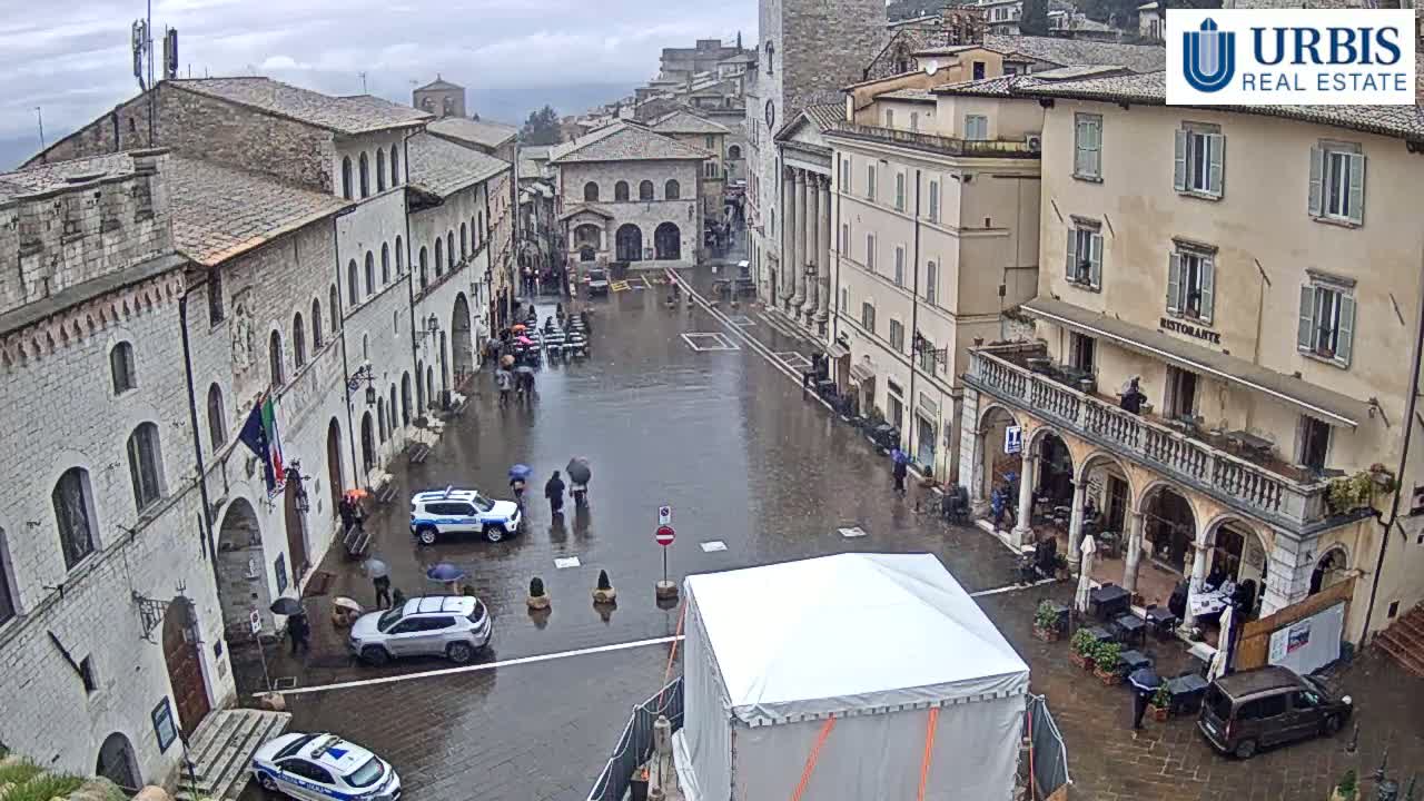 A rain-soaked town square in a European-style town, with several cars parked and people walking around under overcast skies.