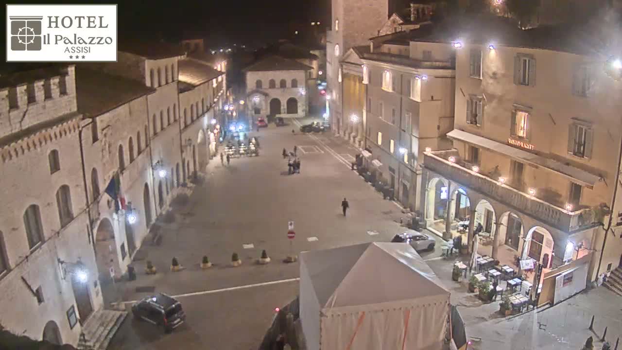 A nighttime, high-angle view of a sparsely populated town square with several buildings illuminated, featuring a large white tent in the foreground.