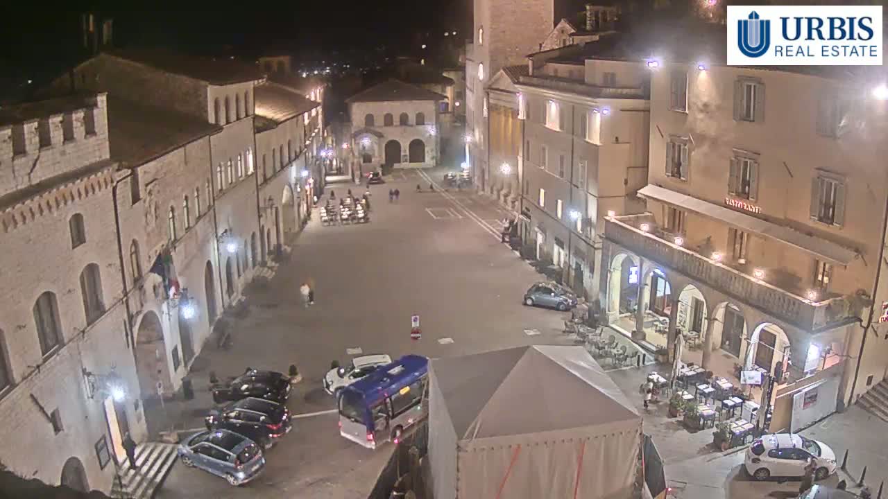 A nighttime, aerial view of a town square surrounded by historic buildings, with cars parked along the edges and a few people visible.