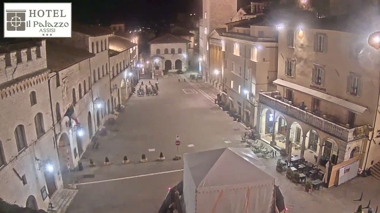 An empty, illuminated town square at night, surrounded by old stone buildings.