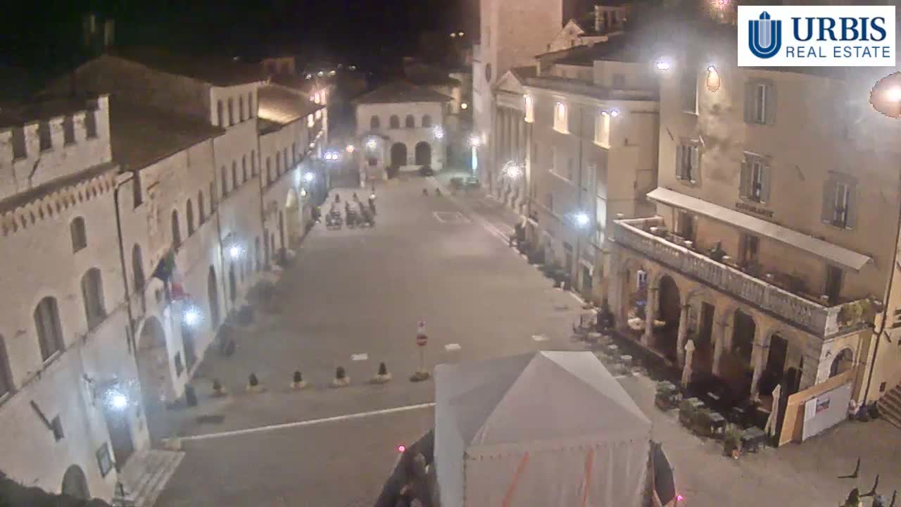 A nighttime, high-angle view of a sparsely populated town square, surrounded by buildings with exterior lighting.