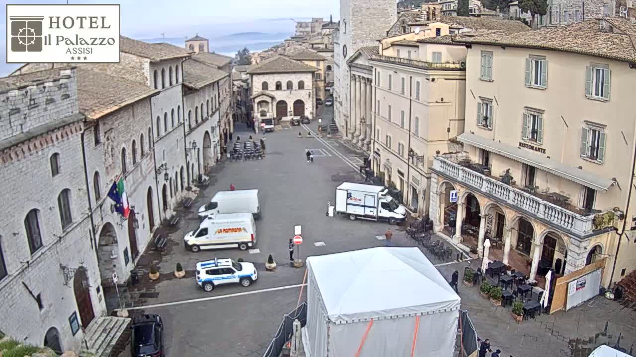 A mostly empty town square in a historic Italian town on an overcast day, with several vehicles, including delivery trucks and police cars, parked around the perimeter.