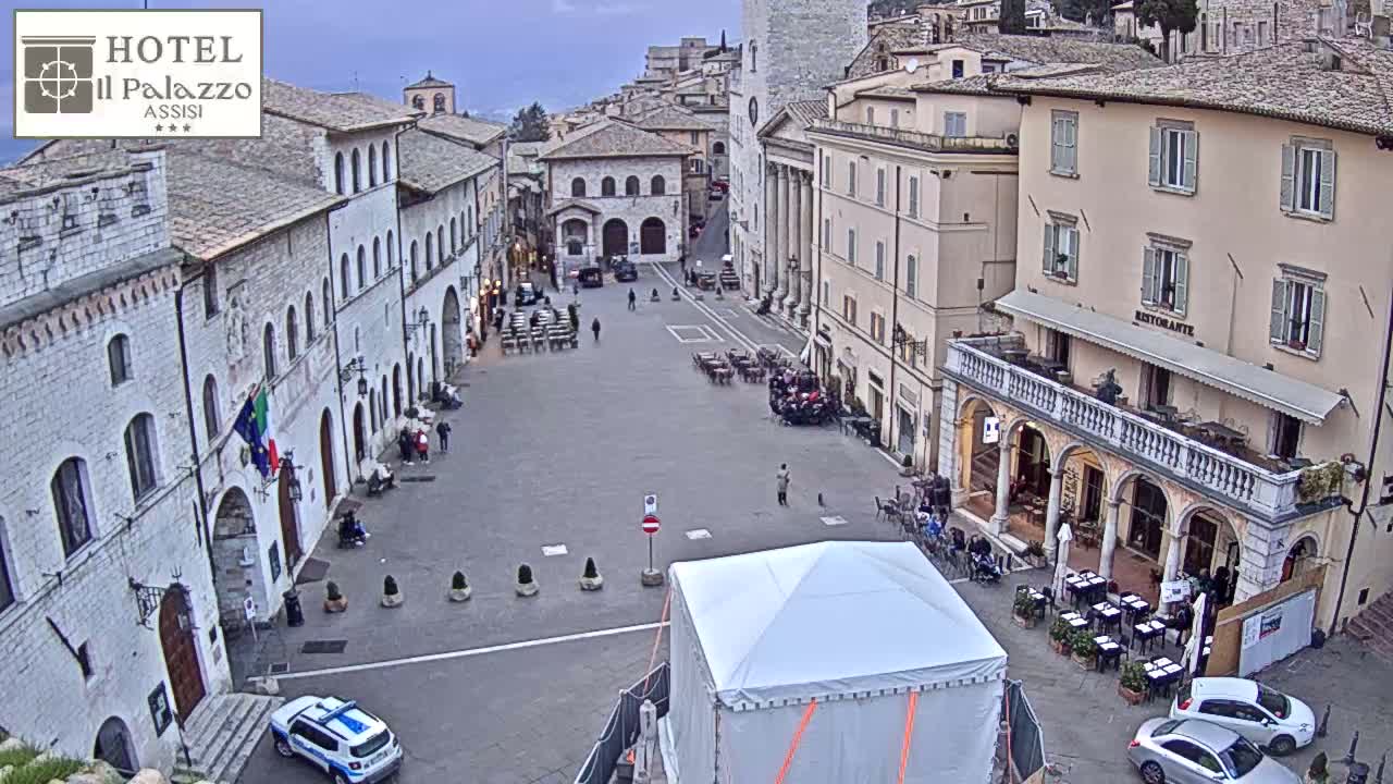 A large, mostly empty town square is surrounded by aged stone buildings under an overcast sky; a white tent sits in the center of the square near a police car and several outdoor cafes.