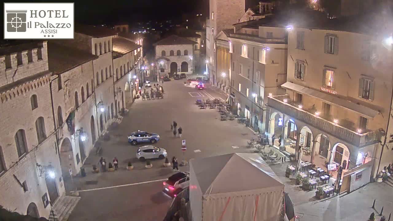 A nighttime, aerial view of a town square in Italy, showing several buildings lit up, cars parked, and a few people walking around.