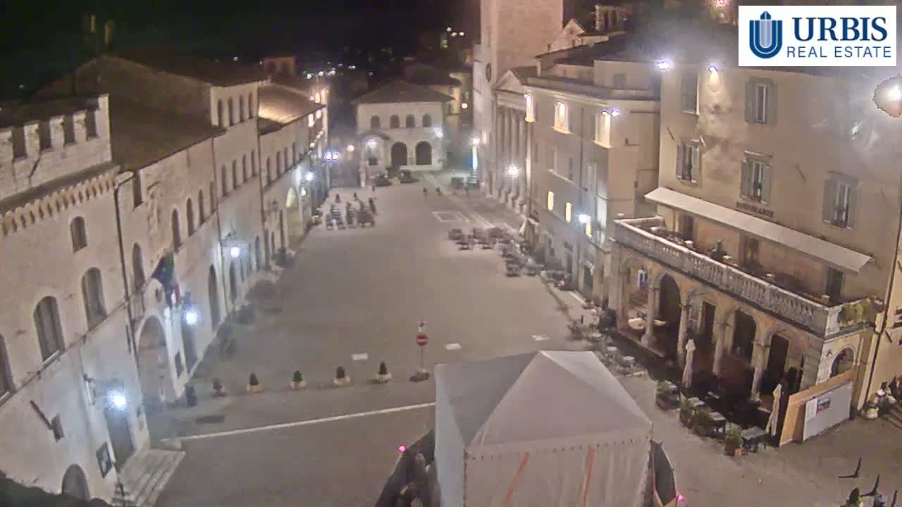 An empty town square at night, surrounded by illuminated buildings, with a few small groups of people and outdoor seating areas.