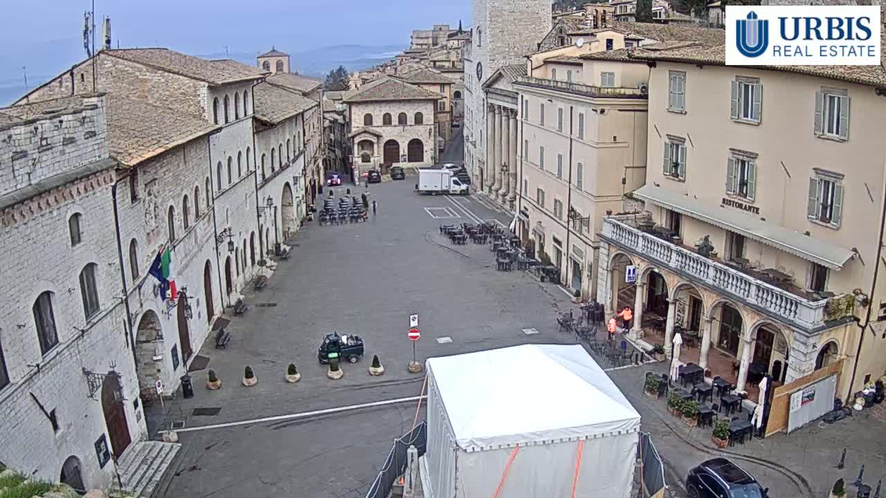 A mostly empty town square in a European-style town on an overcast day, with several buildings surrounding it, some outdoor seating, and a large white tent in the center.