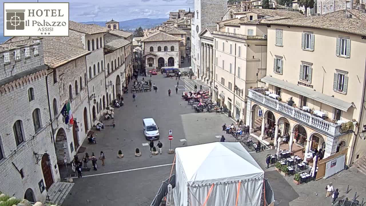 A mostly sunny day reveals a large town square with several buildings, outdoor seating areas, and a few people milling about; a white tent sits in the center.
