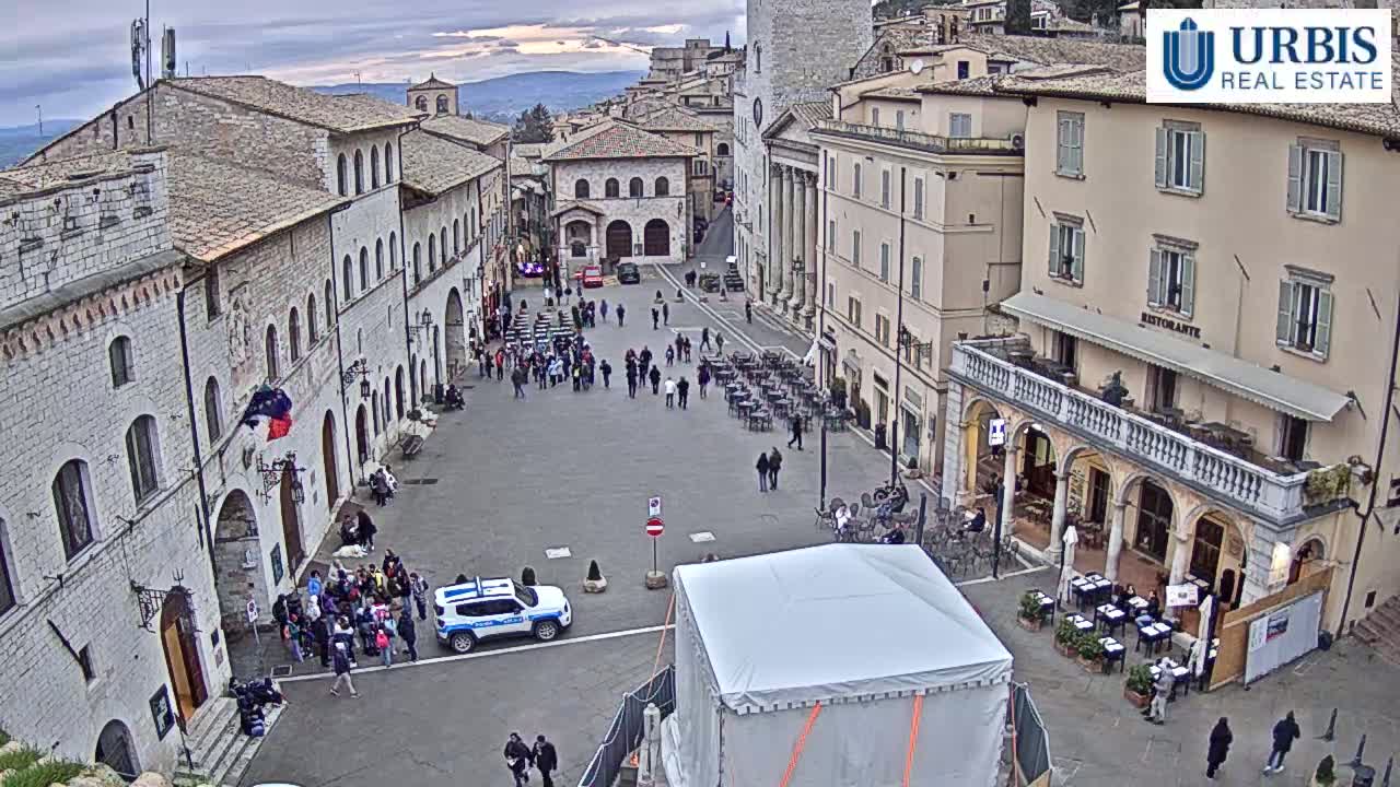 A large, mostly empty town square with several buildings, outdoor seating areas, and a few people, under an overcast sky.