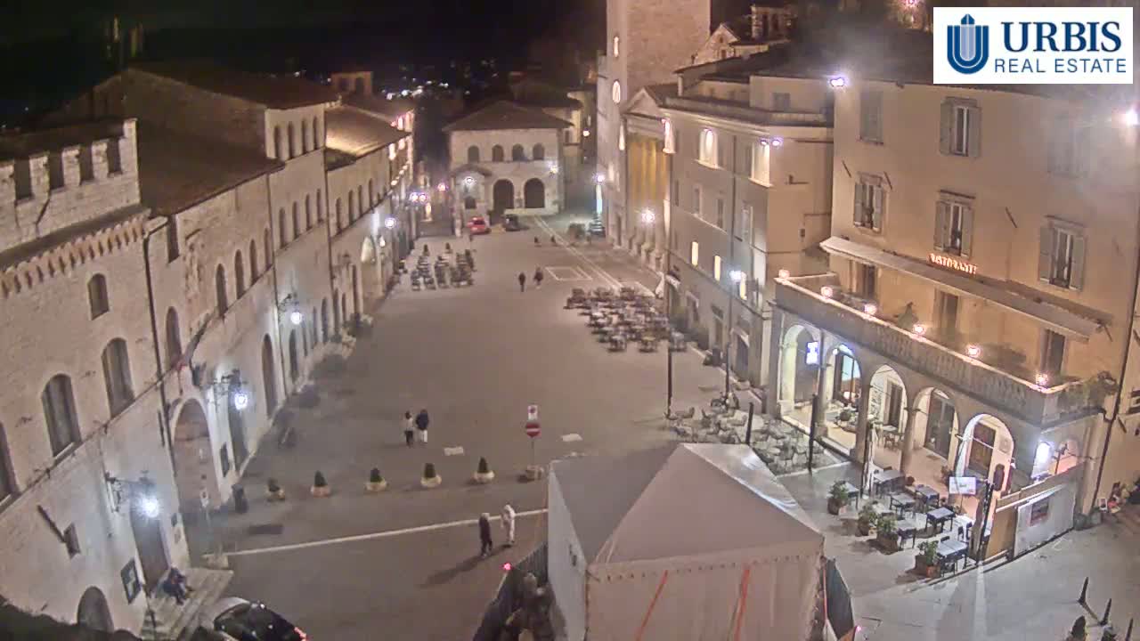 A nighttime view of a town square surrounded by illuminated buildings, with a few people and outdoor seating areas visible.