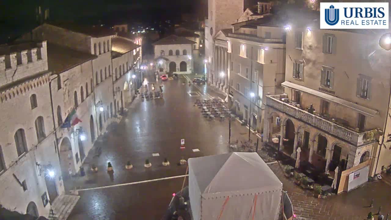 A wet, empty town square at night, surrounded by old stone buildings with lights illuminating the scene.