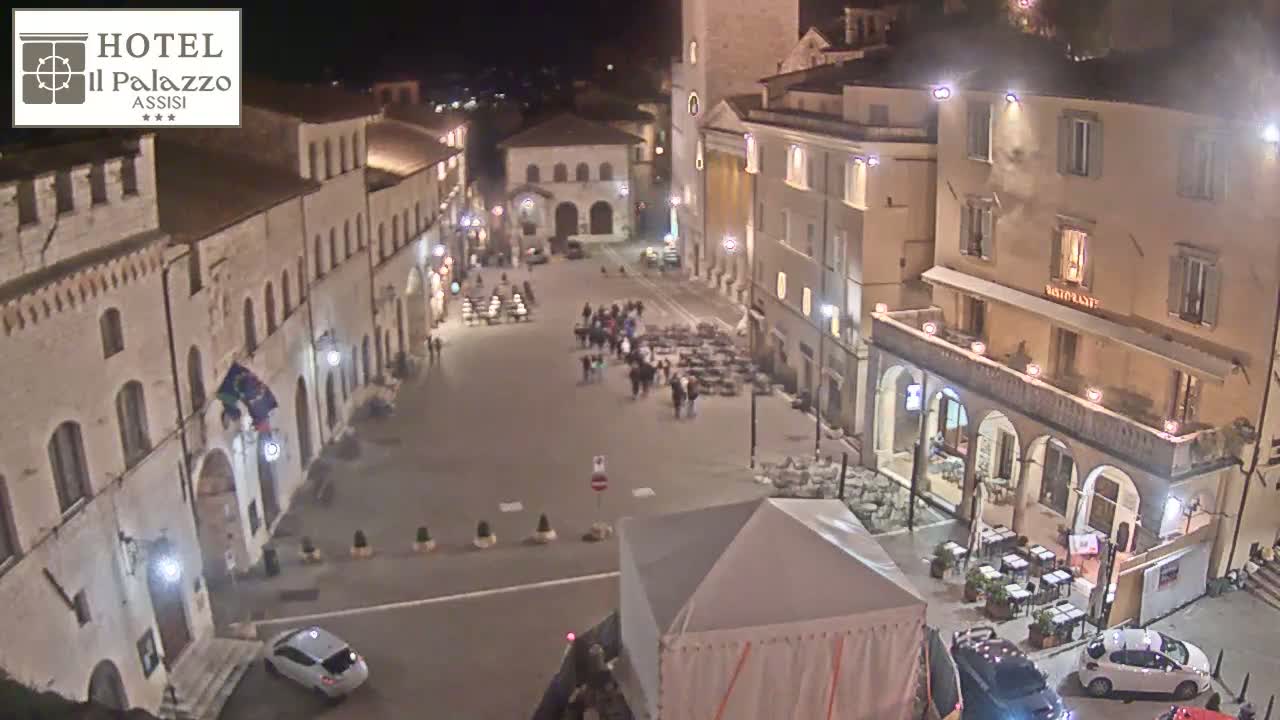 A nighttime, high-angle view of a town square with several buildings illuminated, people walking, and outdoor seating at restaurants.
