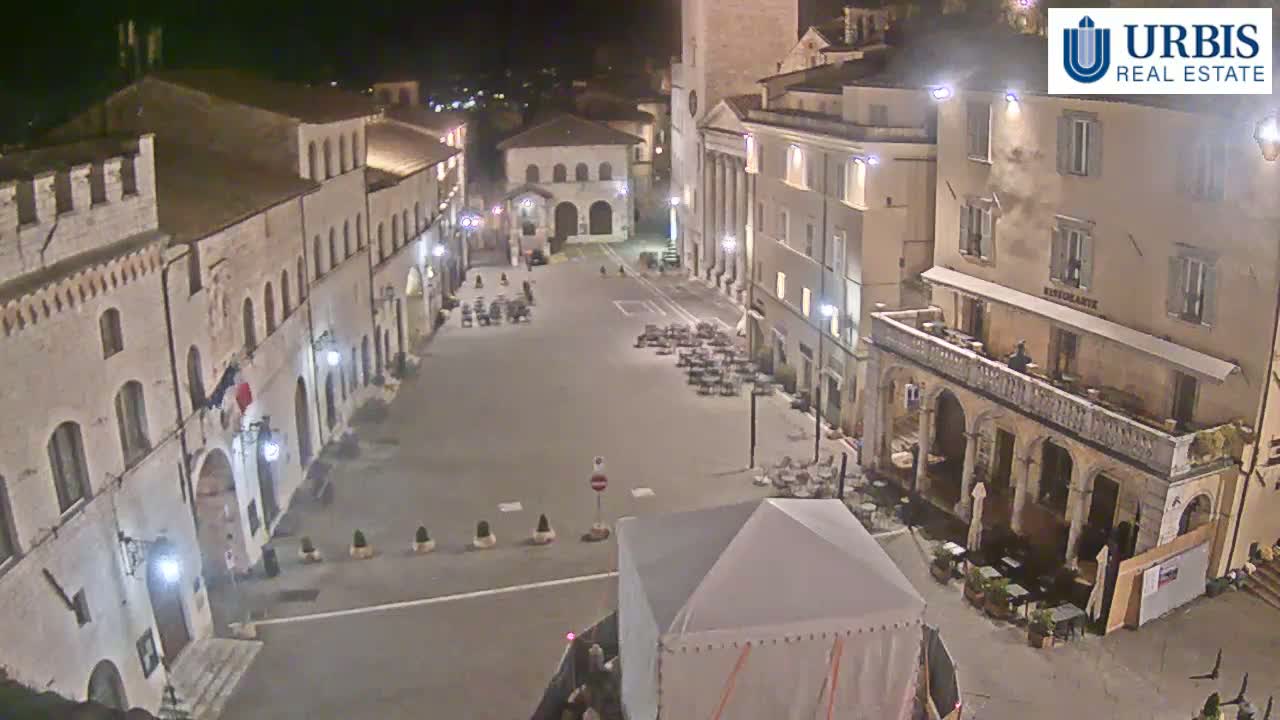 A nighttime view of a mostly empty town square surrounded by historic-looking buildings, with a few outdoor café tables and chairs visible and a large tent in the foreground.