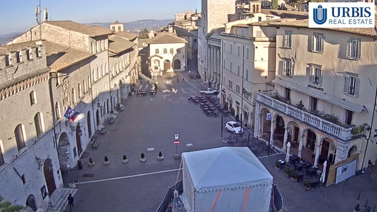 A large town square with stone buildings surrounding it, a few cars parked, and outdoor seating at a restaurant, on a sunny day.