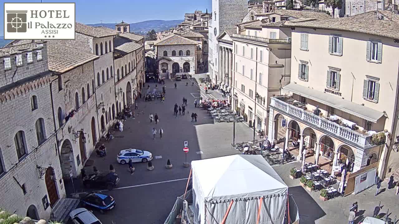 A sunny town square in Italy, with several people walking around, cars parked, and outdoor seating at a restaurant, as well as a large white tent in the center.