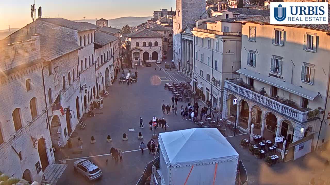 A sunlit town square with several buildings, outdoor seating areas, pedestrians, and a large white tent.
