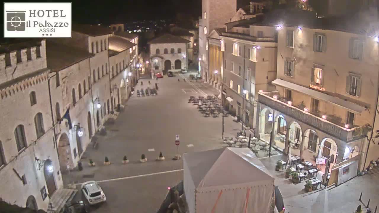 A nighttime, high-angle view of a town square with several buildings, some outdoor seating, and a few cars parked.