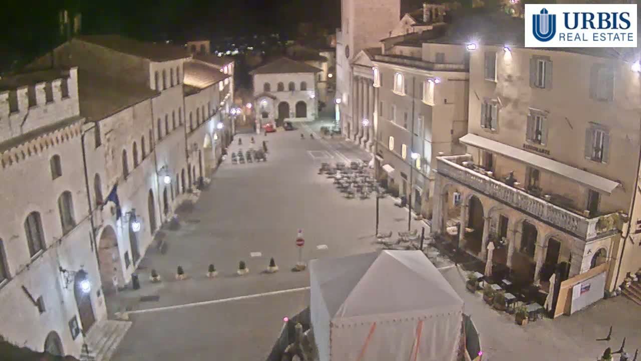 A nighttime view of a town square surrounded by historical buildings, with outdoor seating areas and a large tent in the center.