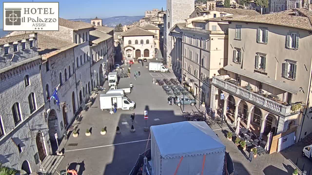 A sunny day reveals a large town square surrounded by historic buildings, with several vans and a few people sparsely scattered throughout.