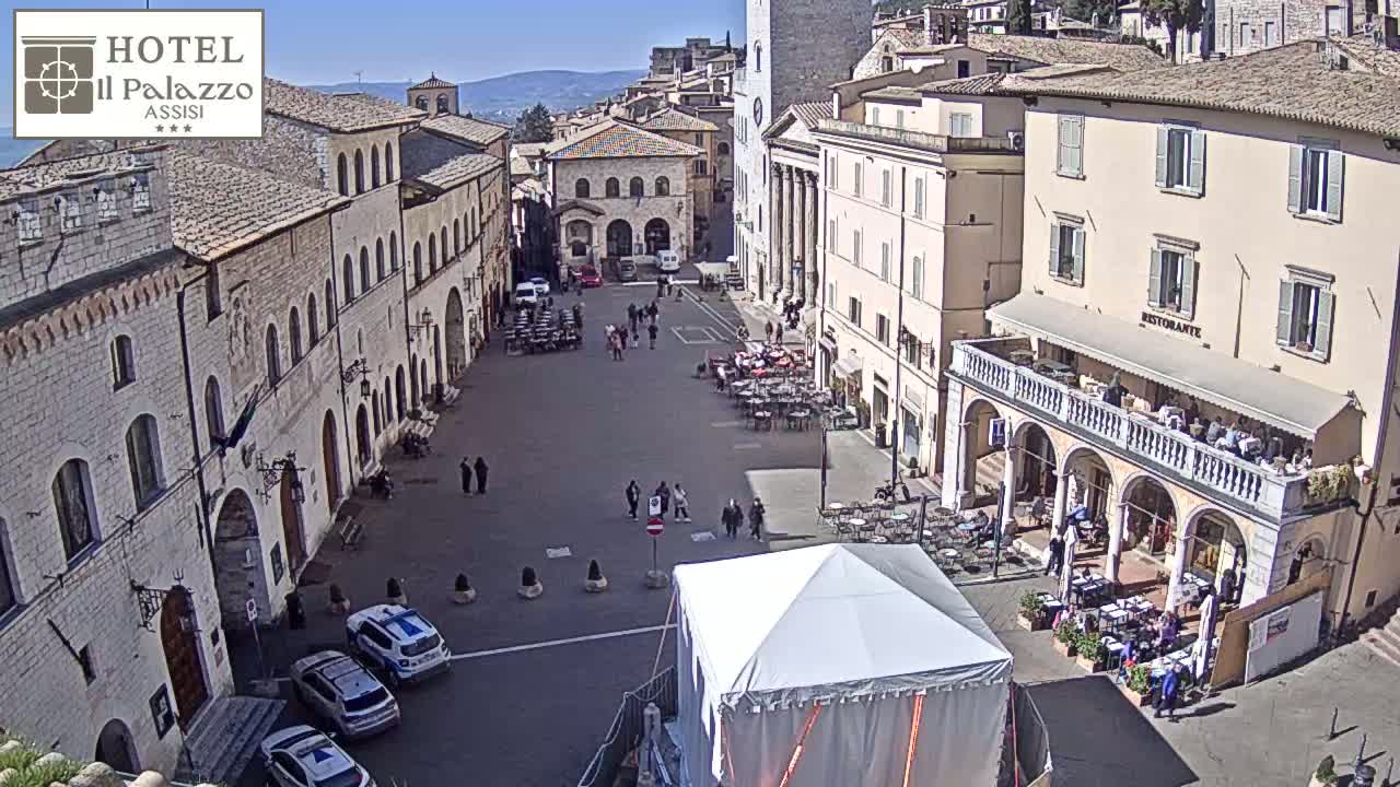 A sunny day in a large European town square with several stone buildings surrounding it, featuring people walking around, outdoor seating at cafes, and police cars parked nearby.