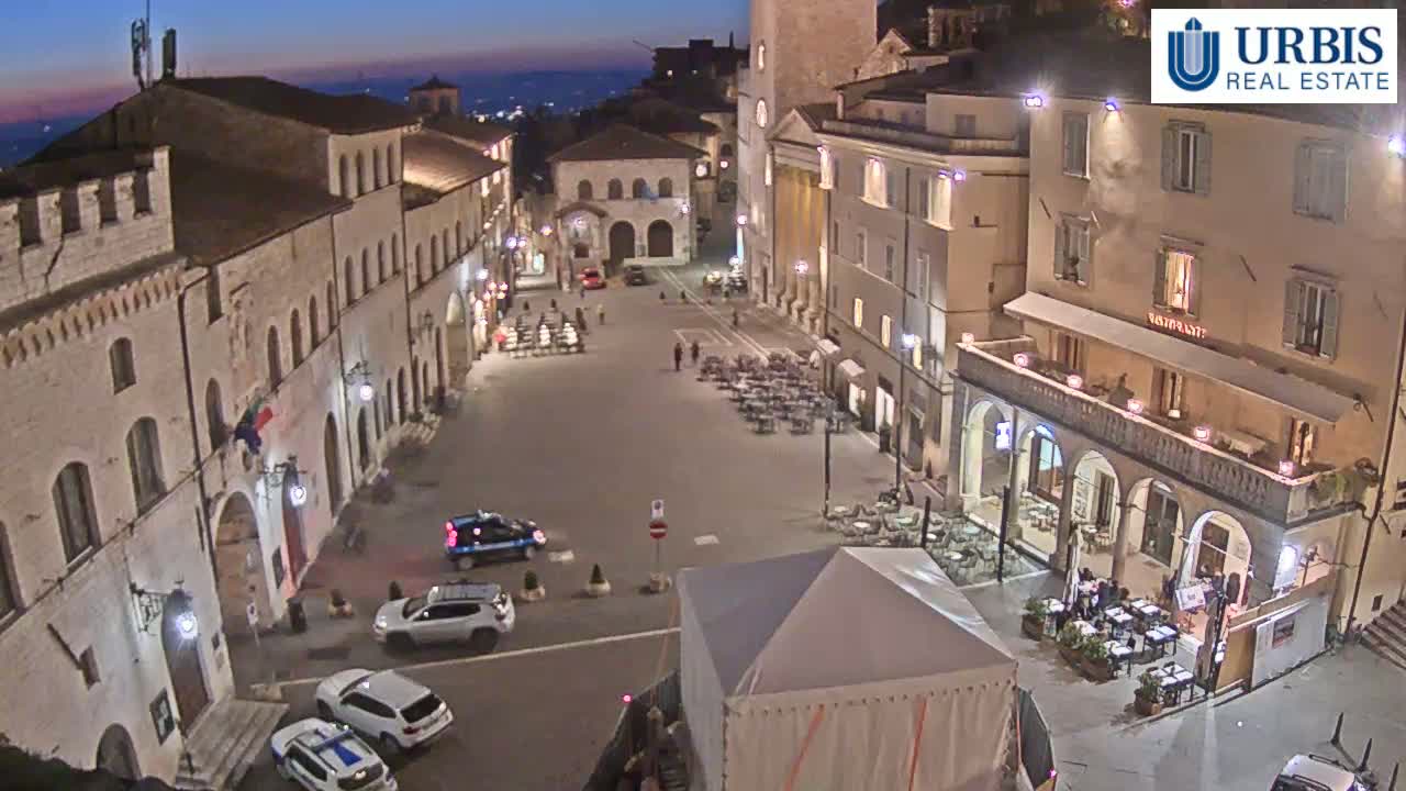 A town square at night, illuminated by streetlights, with several cars parked and outdoor seating areas at restaurants.