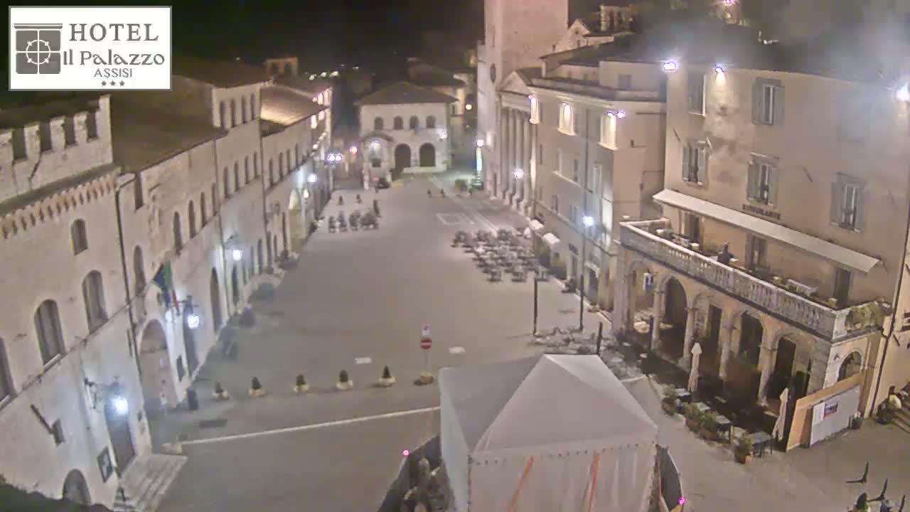 A nighttime view of a large, mostly empty town square surrounded by historic-looking buildings, with a few people and some outdoor seating visible.