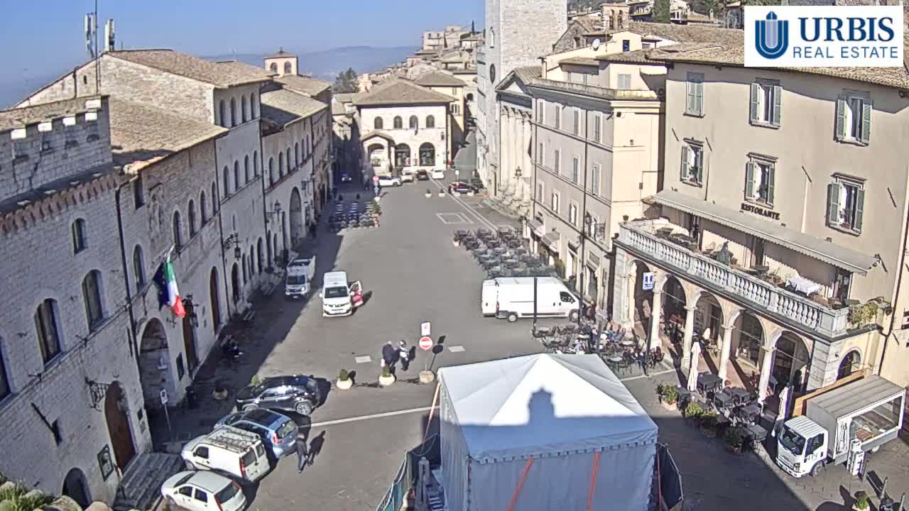 A sunny day in a town square surrounded by historic buildings, with several vehicles parked and some people walking around.
