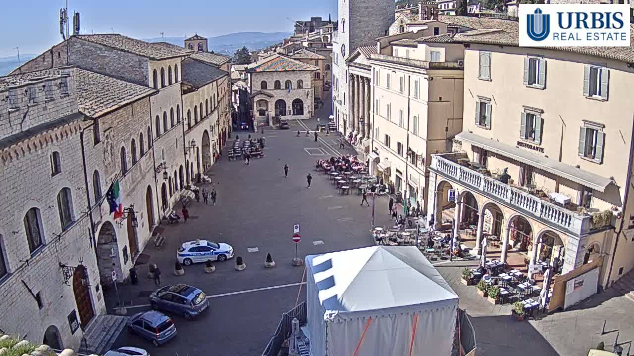 A sunny day in a town square surrounded by historic stone buildings, with cars parked and people seated at outdoor cafes.