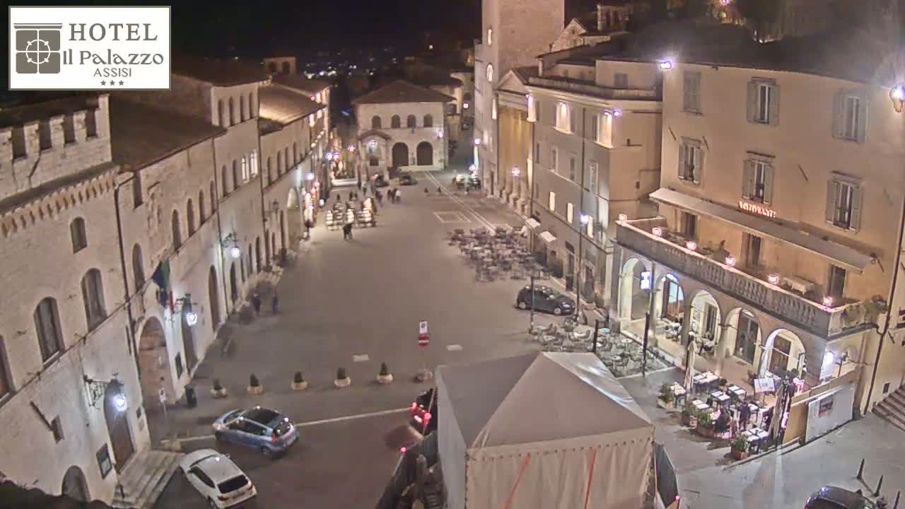 A nighttime, high-angle view of a town square with several buildings, cars, and outdoor seating areas.