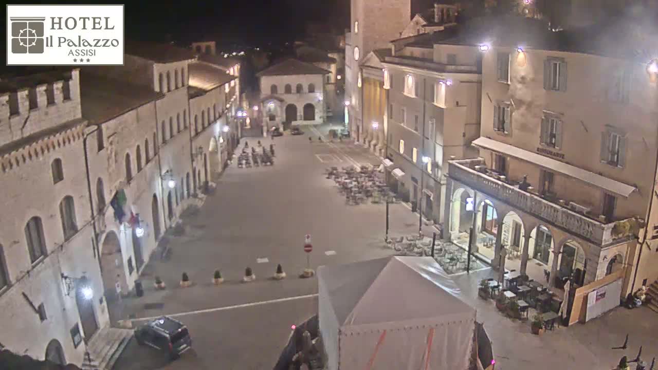A nighttime, high-angle view of a town square in Italy, showing several buildings and sparsely populated outdoor seating areas.