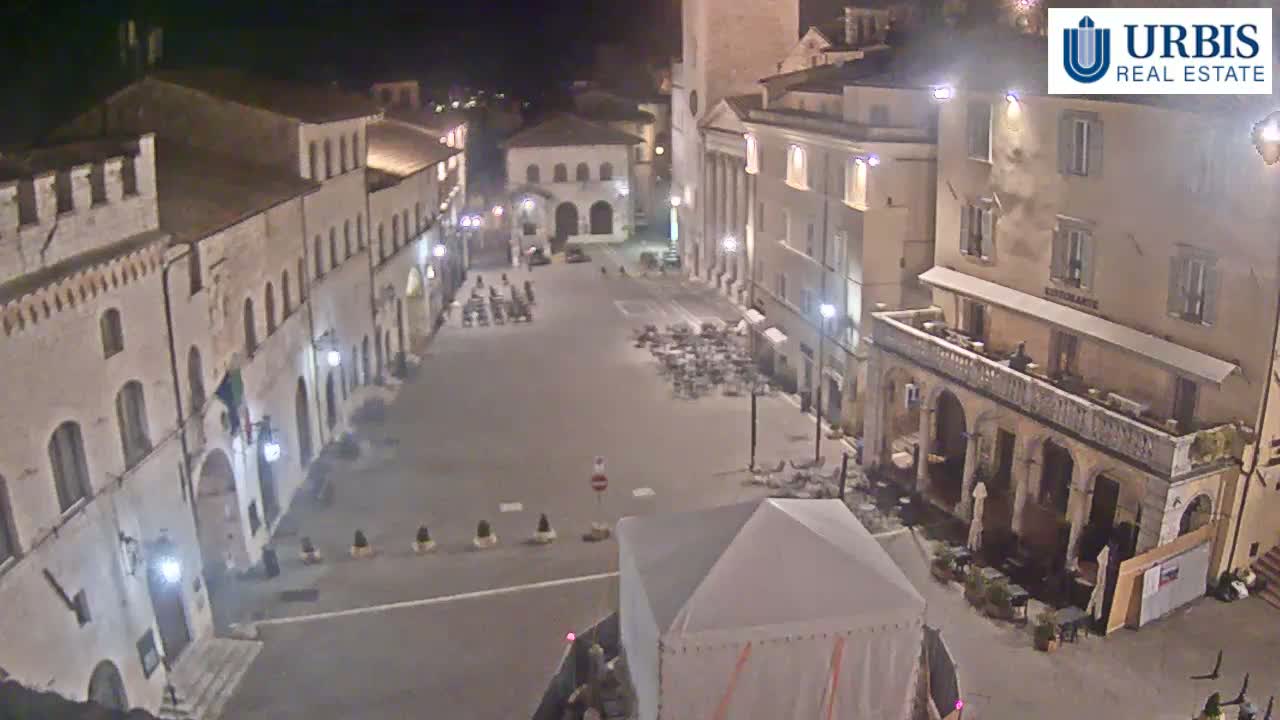 An empty town square at night, surrounded by buildings, with a few chairs and tables scattered about.