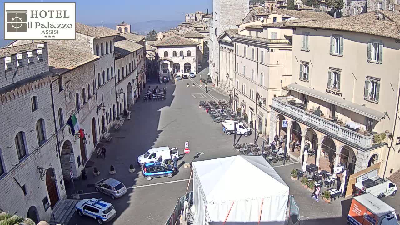 A sunny day in a large, mostly empty town square surrounded by historic buildings, with several cars and a few people present.