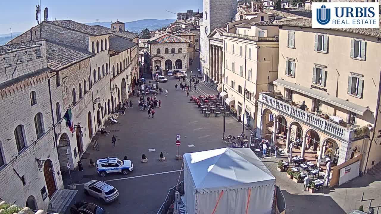 A town square in a sunny European town features several stone buildings, outdoor seating, a few cars, and a large white tent.
