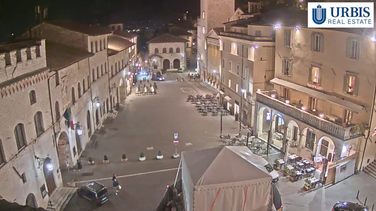 A nighttime, high-angle view of a town square in Italy, surrounded by historical buildings, with several outdoor restaurant seating areas and a few cars parked.