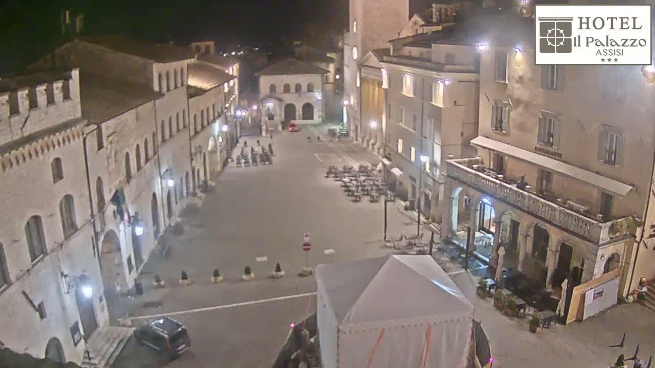 A nighttime, high-angle view of a town square with several buildings surrounding it, sparsely populated with outdoor furniture and a few people.