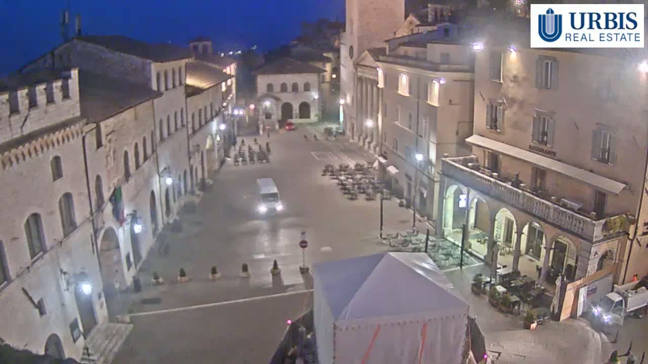 A mostly empty town square at night, under dark, clear skies, is surrounded by tall buildings with outdoor seating areas.