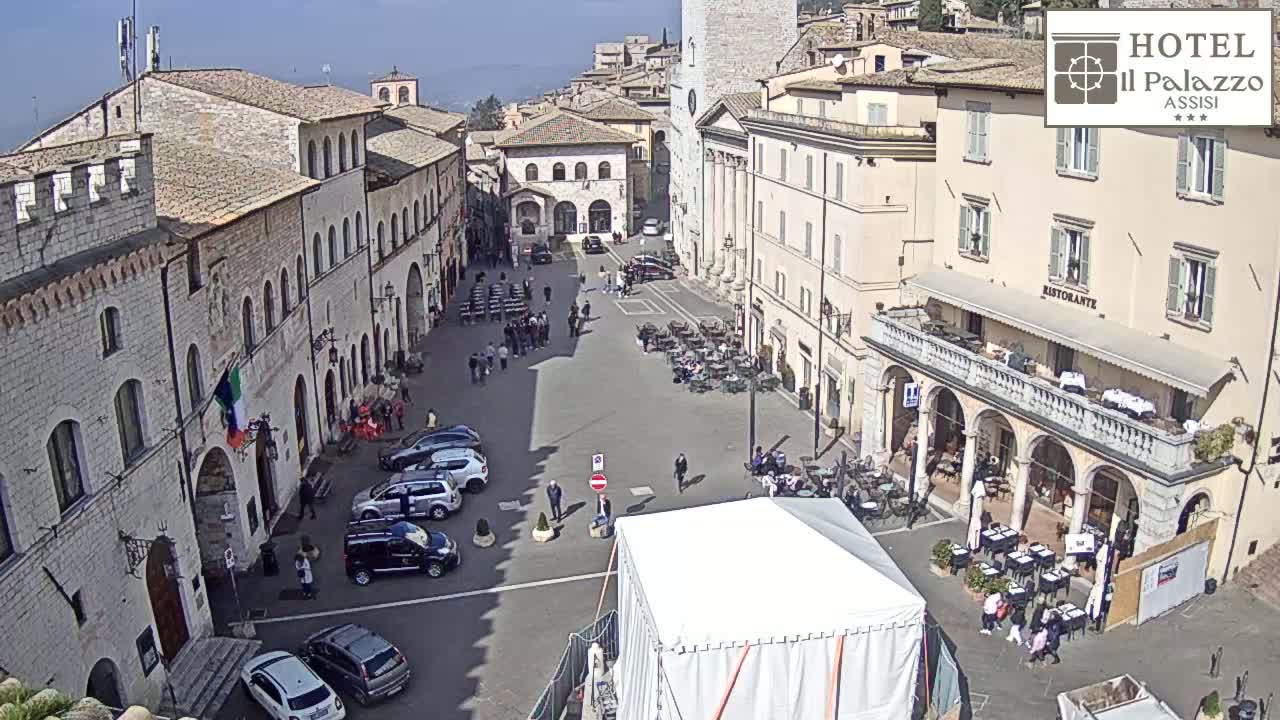 A sunny day reveals a large town square in Italy, surrounded by ancient stone buildings, with cars parked and people sitting at outdoor cafes.