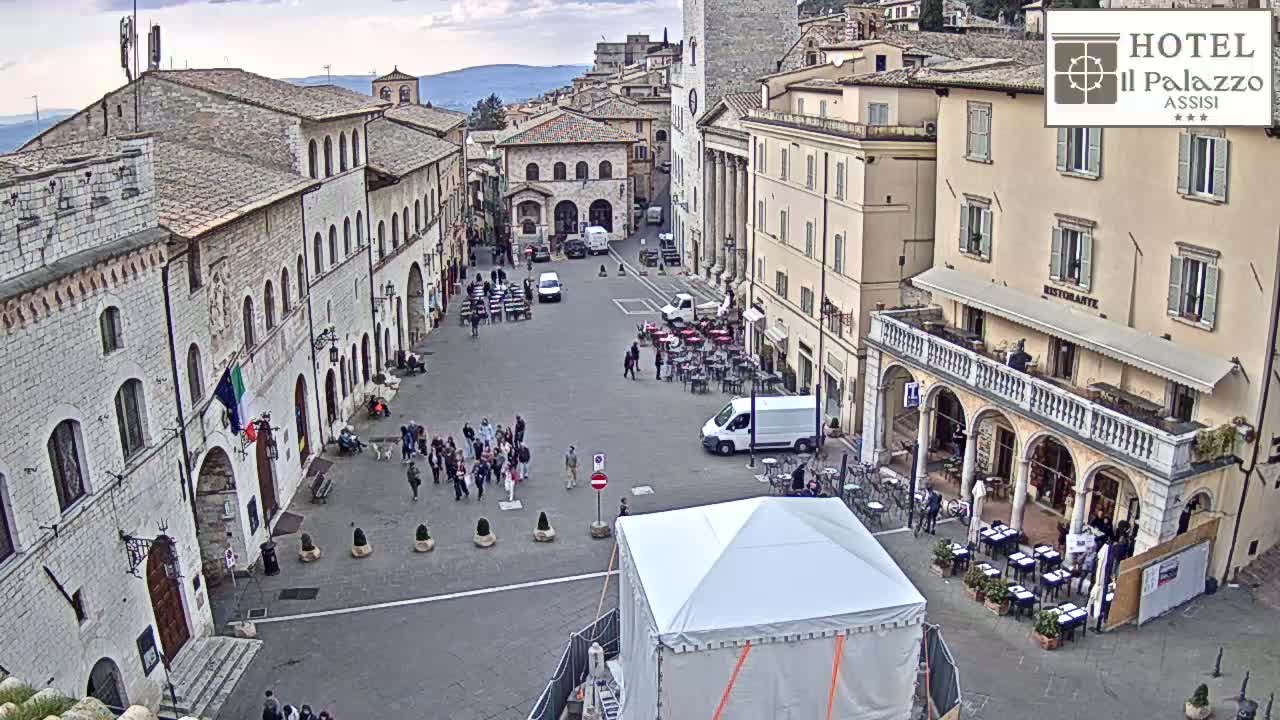 A mostly empty town square in an overcast setting is surrounded by multiple-story stone buildings, with several people walking around and some outdoor seating at a restaurant.