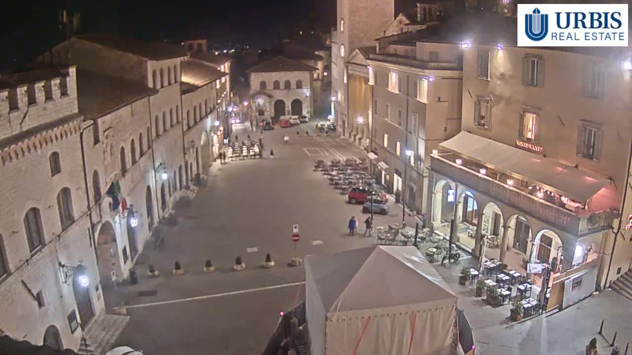 A nighttime, high-angle view of a mostly empty town square in a European-style town, with several buildings surrounding it and outdoor seating at a restaurant.