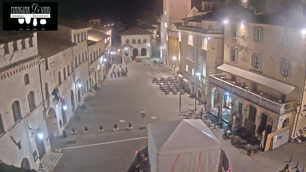 A nighttime, high-angle view of a town square in Italy, showing several buildings with outdoor seating areas sparsely populated under the streetlights.