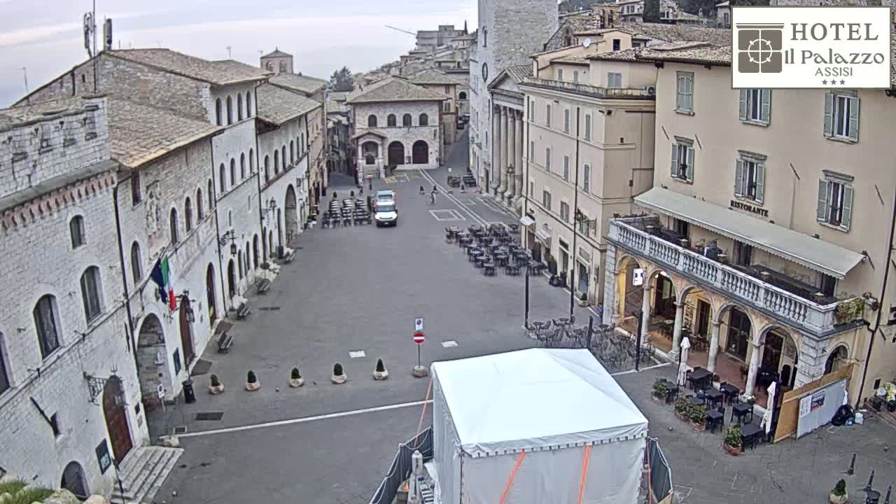 A mostly empty town square in overcast weather is surrounded by ancient stone buildings, with some outdoor seating and a covered structure in the center.