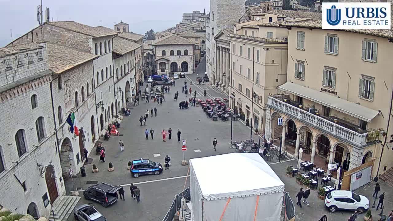 A large, paved town square is surrounded by stone buildings under an overcast sky, with several people walking around and cars parked.