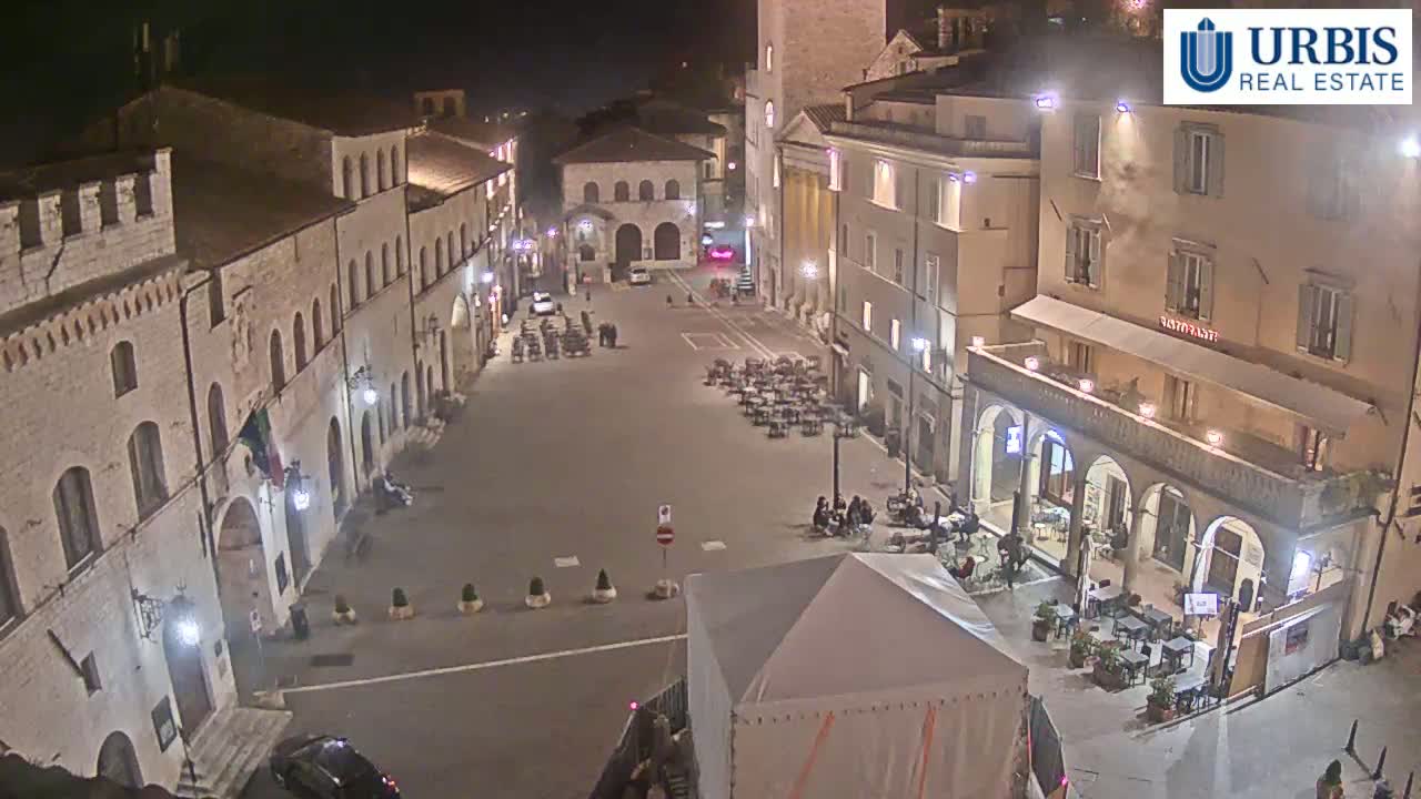 A nighttime, high-angle view of a town square in Italy, featuring stone buildings with outdoor seating areas and a few people present.